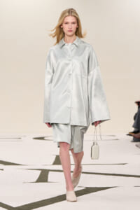 Female model walks a runway in a loose, silver satin shirt and matching shorts, carrying a small pale bag and wearing beige mules.