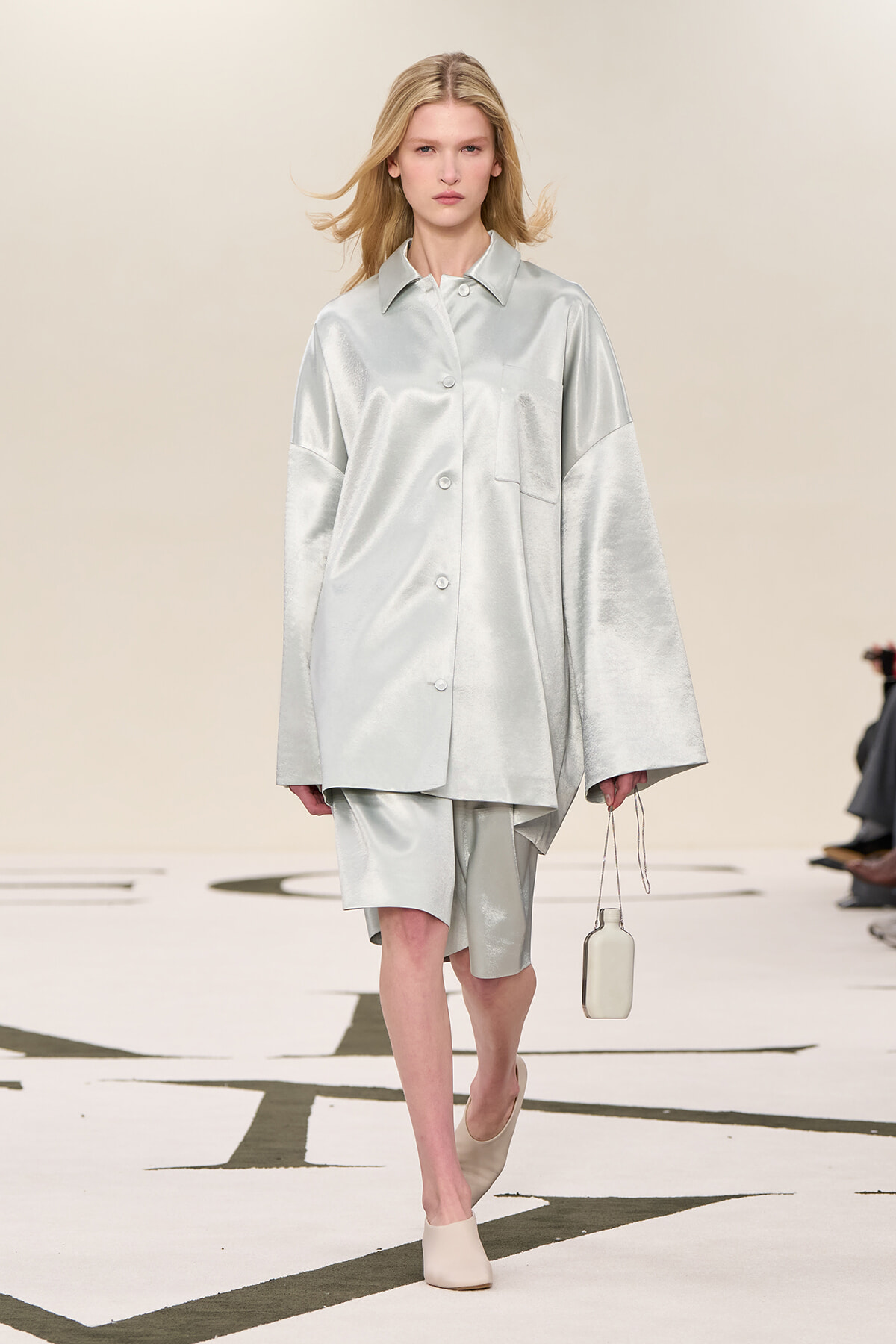 Female model walks a runway in a loose, silver satin shirt and matching shorts, carrying a small pale bag and wearing beige mules.