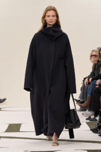 Model walking a fashion show in a long, oversized black coat with a high, draped collar, carrying a black bucket bag and wearing beige flats. Audience members seated along the runway are visible in the background.