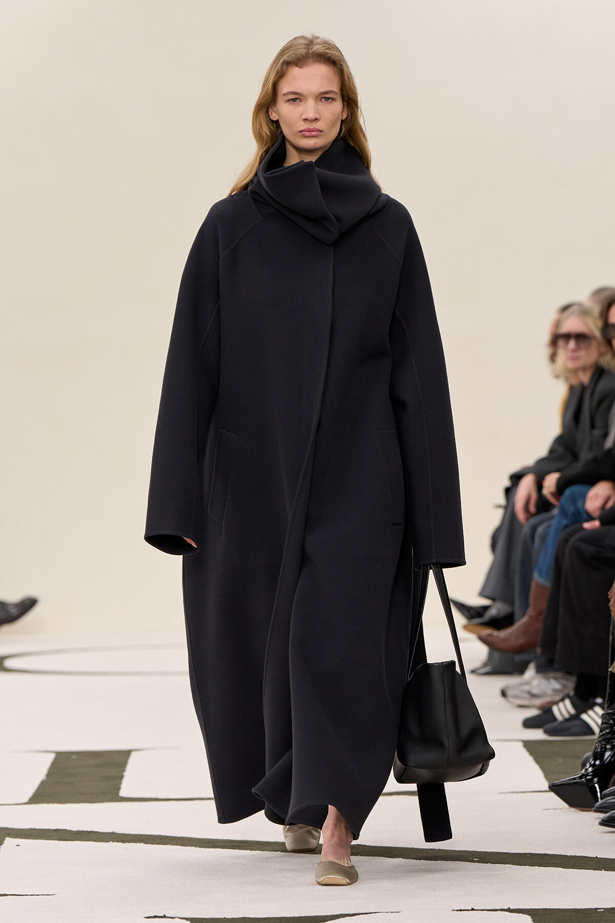 Model walking a fashion show in a long, oversized black coat with a high, draped collar, carrying a black bucket bag and wearing beige flats. Audience members seated along the runway are visible in the background.