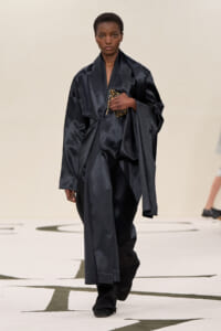 Model walks runway in an oversized black satin ensemble, holding a small leopard-print clutch at chest height?