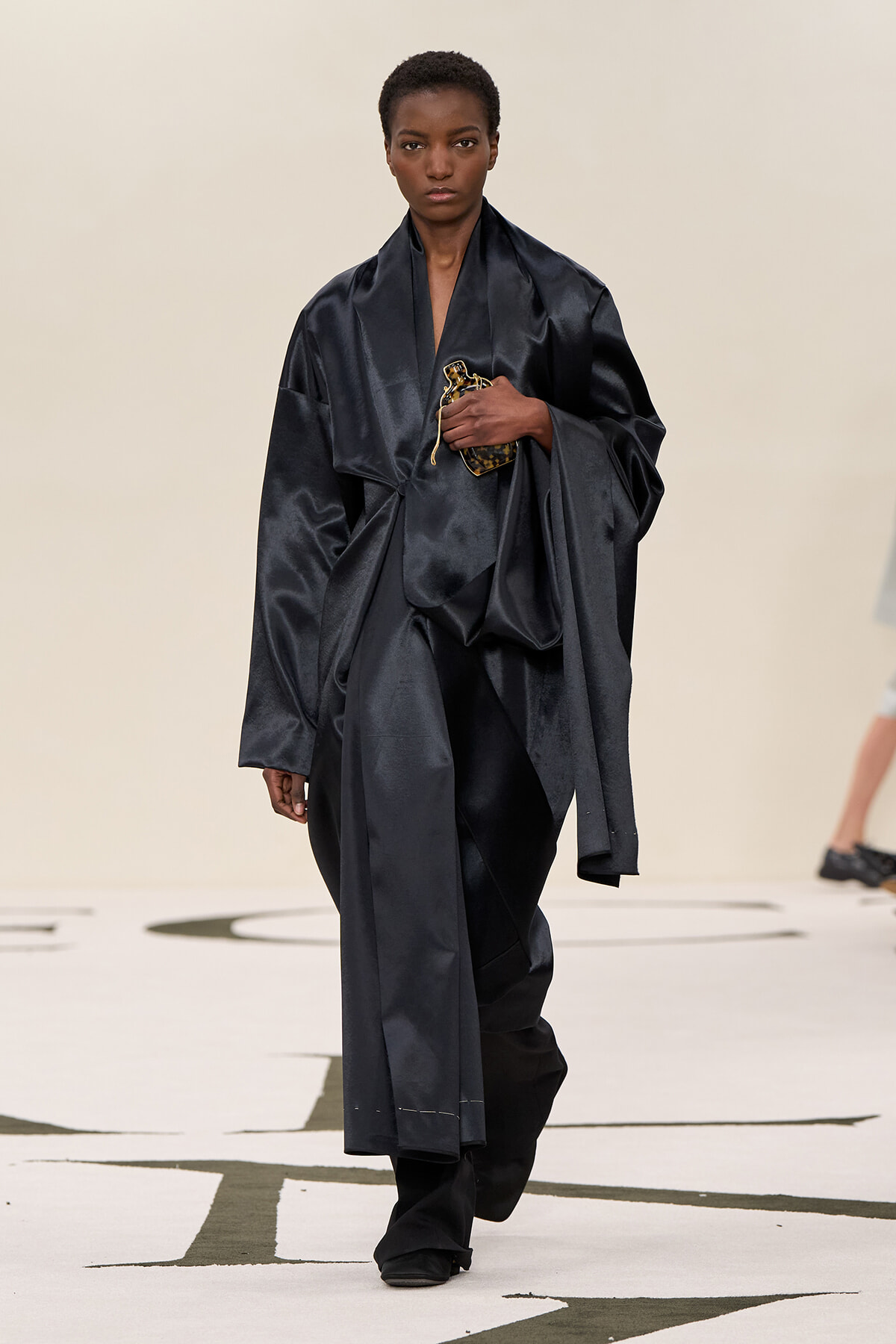 Model walks runway in an oversized black satin ensemble, holding a small leopard-print clutch at chest height?