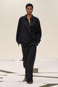 Model wearing an oversized black suit walking on a runway, with a loose shirt-front and relaxed pants.