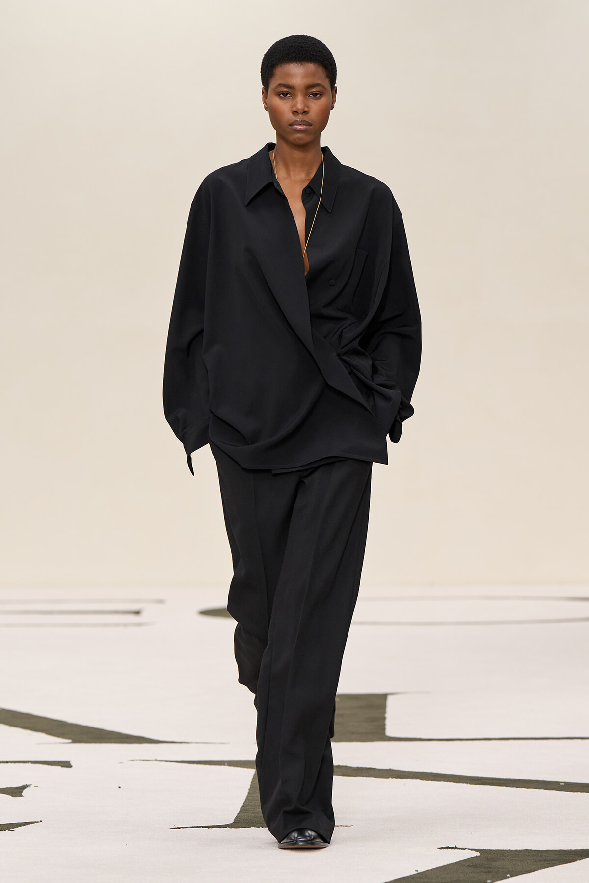 Model wearing an oversized black suit walking on a runway, with a loose shirt-front and relaxed pants.