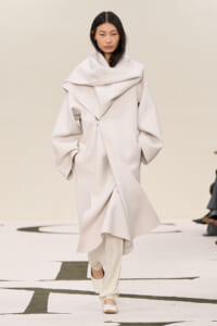 Model walking a runway in an oversized pale beige coat with a large draped collar and wrap details.