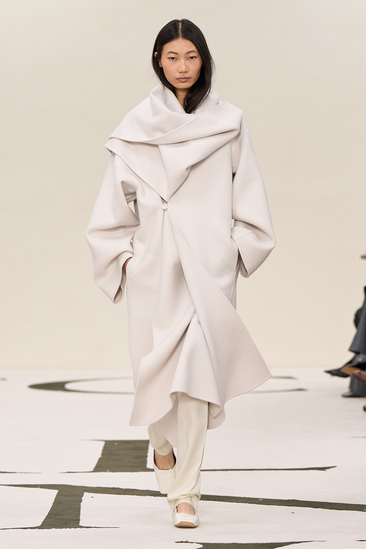 Model walking a runway in an oversized pale beige coat with a large draped collar and wrap details.