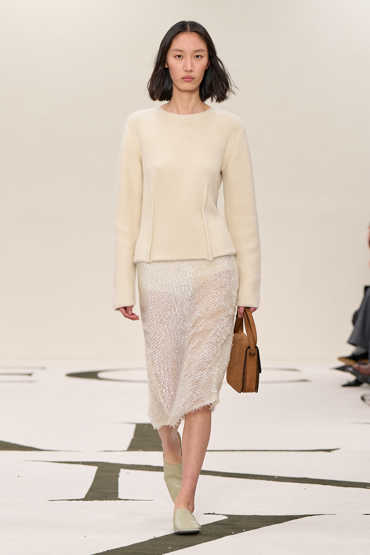 Full-body shot of a model walking a runway in a cream knit sweater and textured midi skirt, carrying a brown handbag.