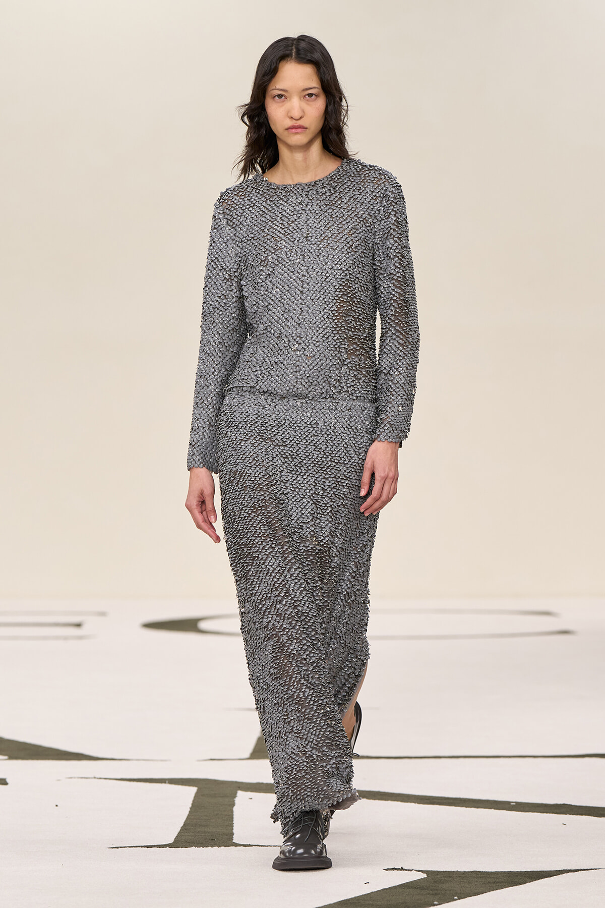Model walking the runway in a textured gray, long-sleeve knit maxi dress with black shoes.