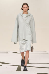 Model walking in a light gray oversized top with a wide, draped collar and matching skirt on a runway, holding a small metallic bag.