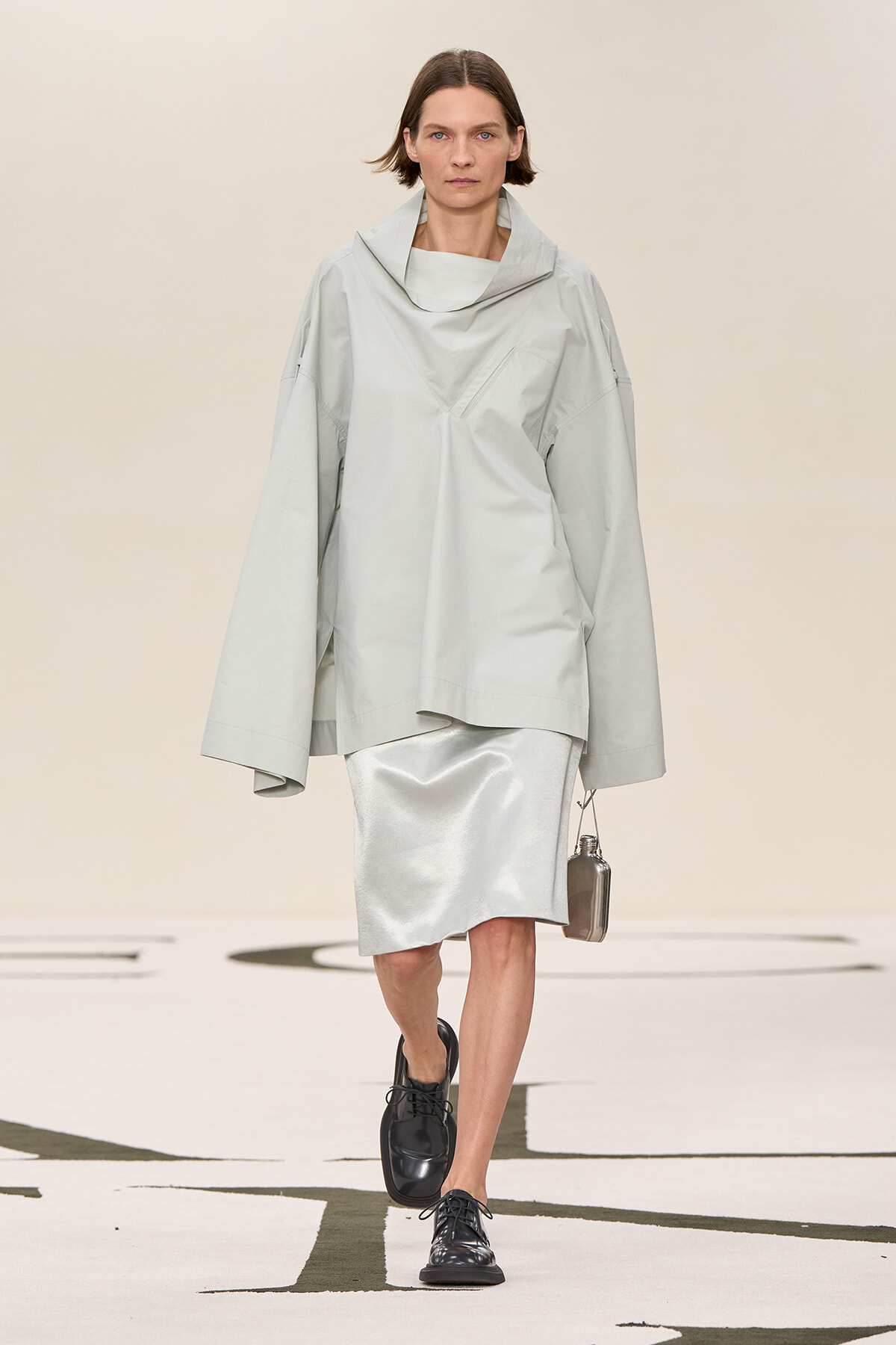 Model walking in a light gray oversized top with a wide, draped collar and matching skirt on a runway, holding a small metallic bag.
