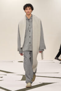 Male model walking a runway in a gray tonal outfit: buttoned shirt, loose pants, and a cream scarf draped over his shoulders.