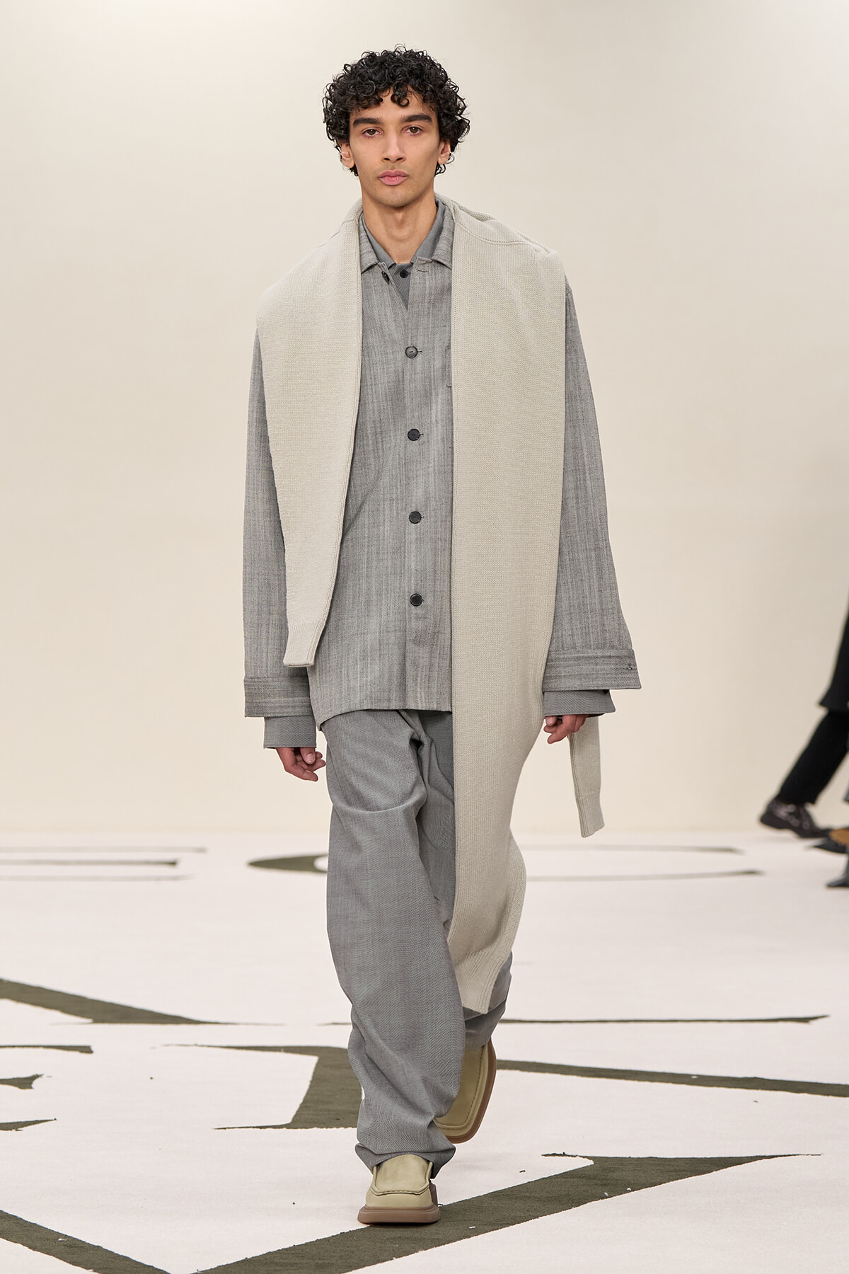 Male model walking a runway in a gray tonal outfit: buttoned shirt, loose pants, and a cream scarf draped over his shoulders.
