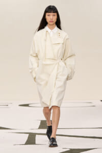 Female model walks a runway in an oversized ivory trench dress with a wrap belt and black oxford shoes.