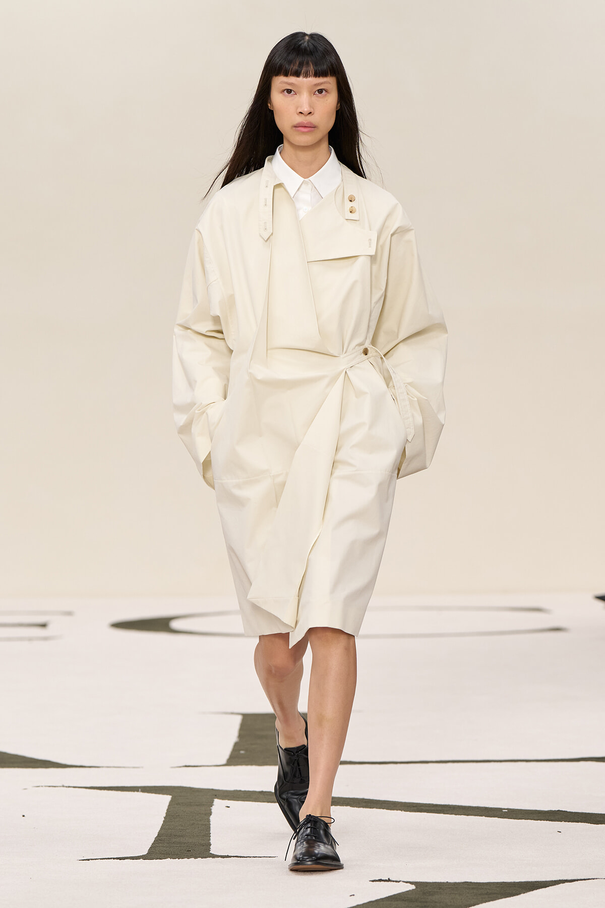 Female model walks a runway in an oversized ivory trench dress with a wrap belt and black oxford shoes.