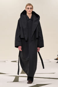 Male model on a runway wearing an oversized black trench with a large hood and sunglasses.