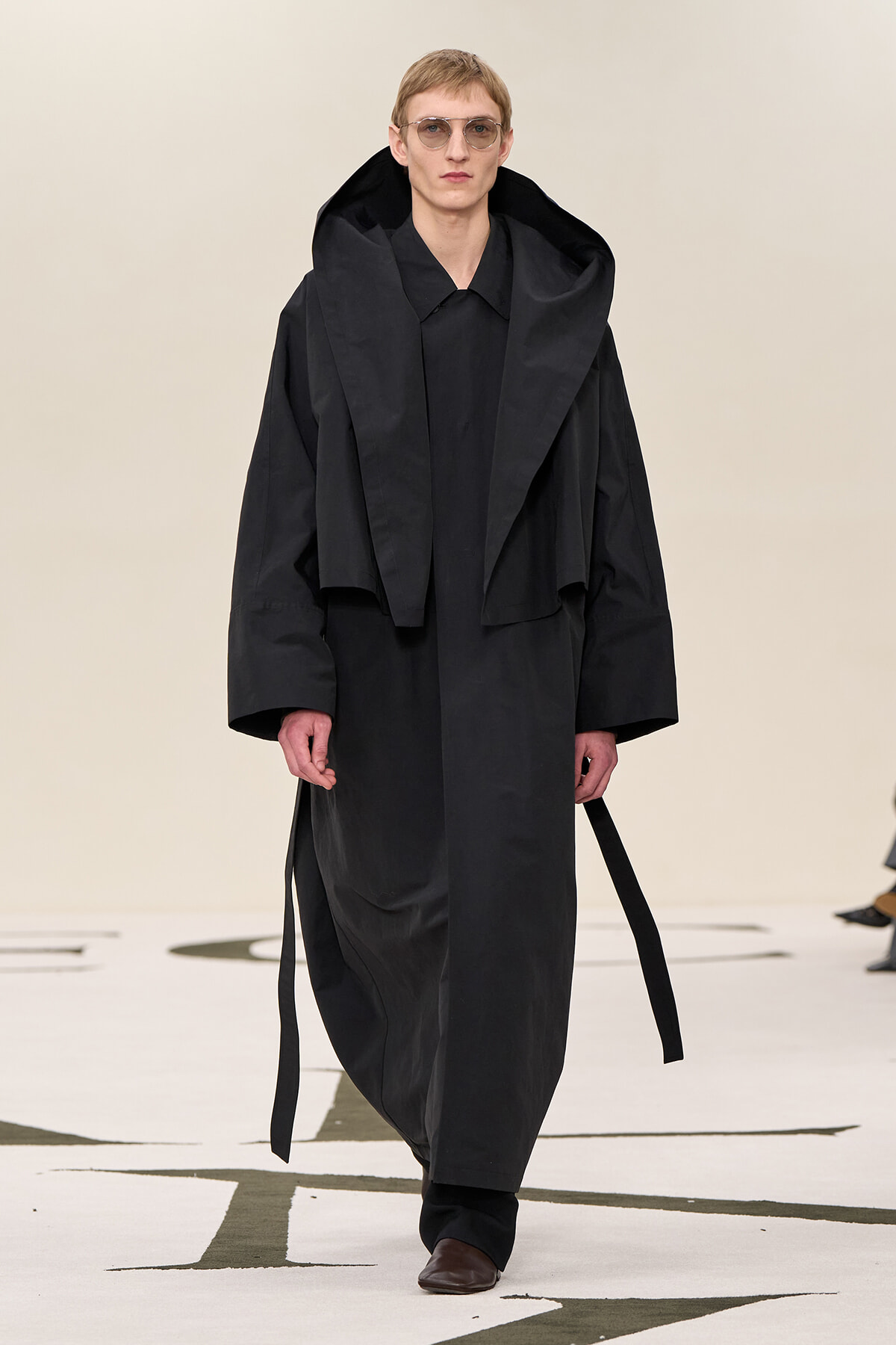 Male model on a runway wearing an oversized black trench with a large hood and sunglasses.