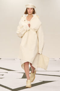 Runway model in an oversized cream coat with a matching hat, beige boots, and a cream tote bag.