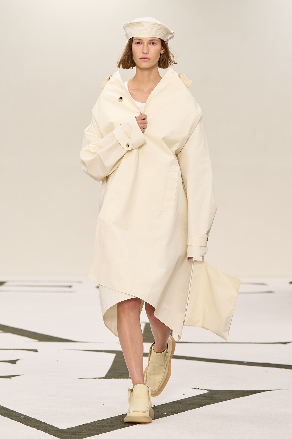 Runway model in an oversized cream coat with a matching hat, beige boots, and a cream tote bag.