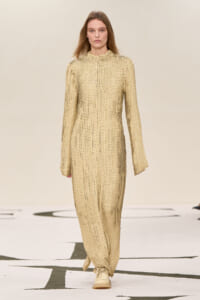 Model walking the runway in a beige, scale-textured, full-length jumpsuit with a high neckline and long sleeves.