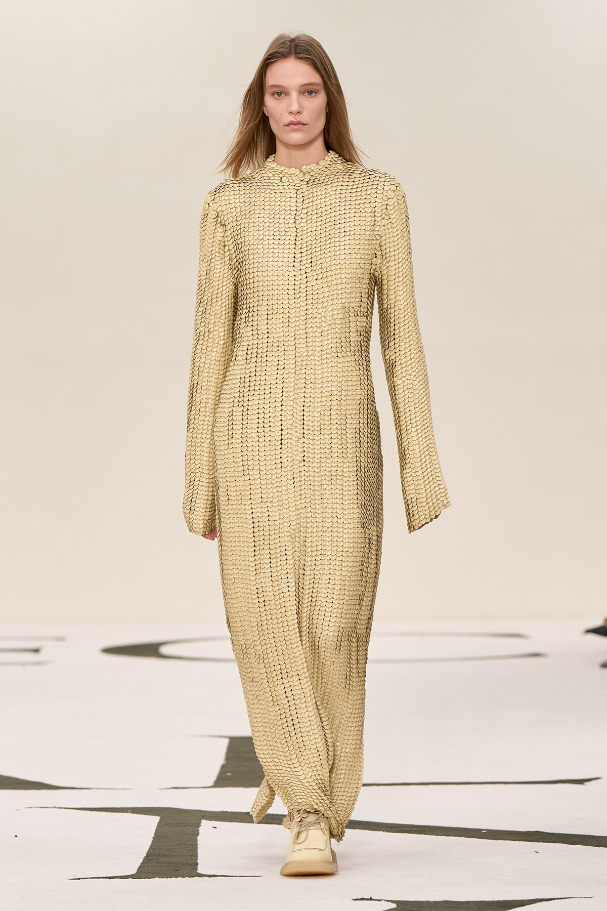 Model walking the runway in a beige, scale-textured, full-length jumpsuit with a high neckline and long sleeves.