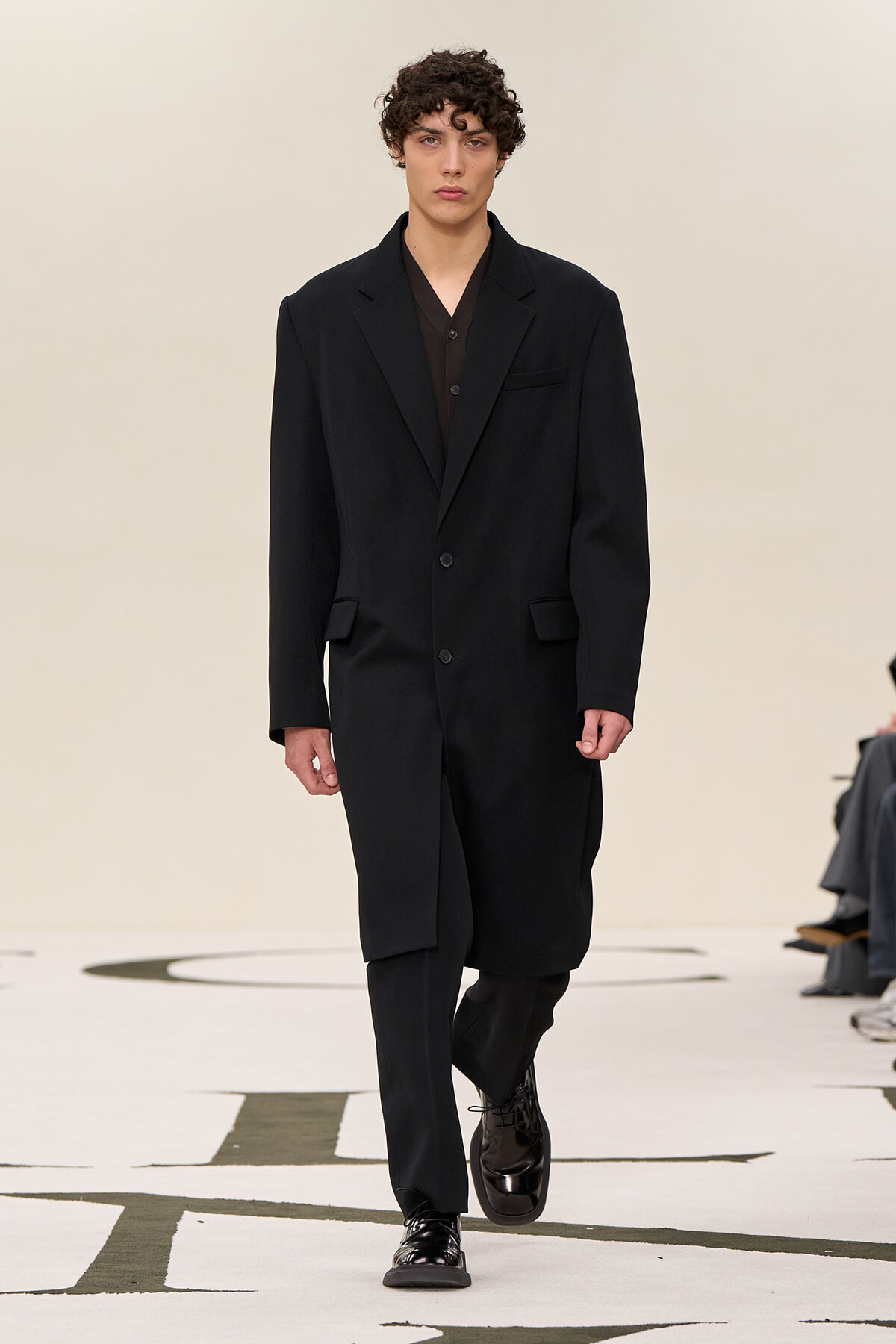 Male model walking a fashion runway in a black oversized suit with a matching dark shirt underneath, neutral expression.