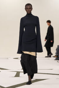 Model on a runway wearing a navy turtleneck top with long flared sleeves and a layered black skirt with a beige underlayer, paired with black flats.