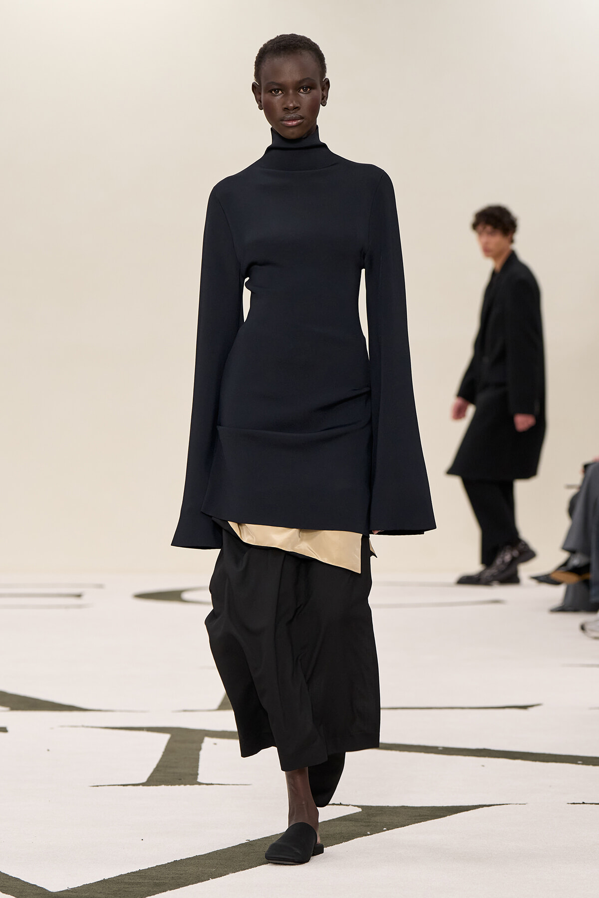 Model on a runway wearing a navy turtleneck top with long flared sleeves and a layered black skirt with a beige underlayer, paired with black flats.