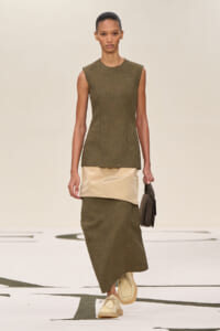 Model wearing an olive-green sleeveless top and long skirt with a beige satin waist panel, carrying a small brown handbag on a runway.