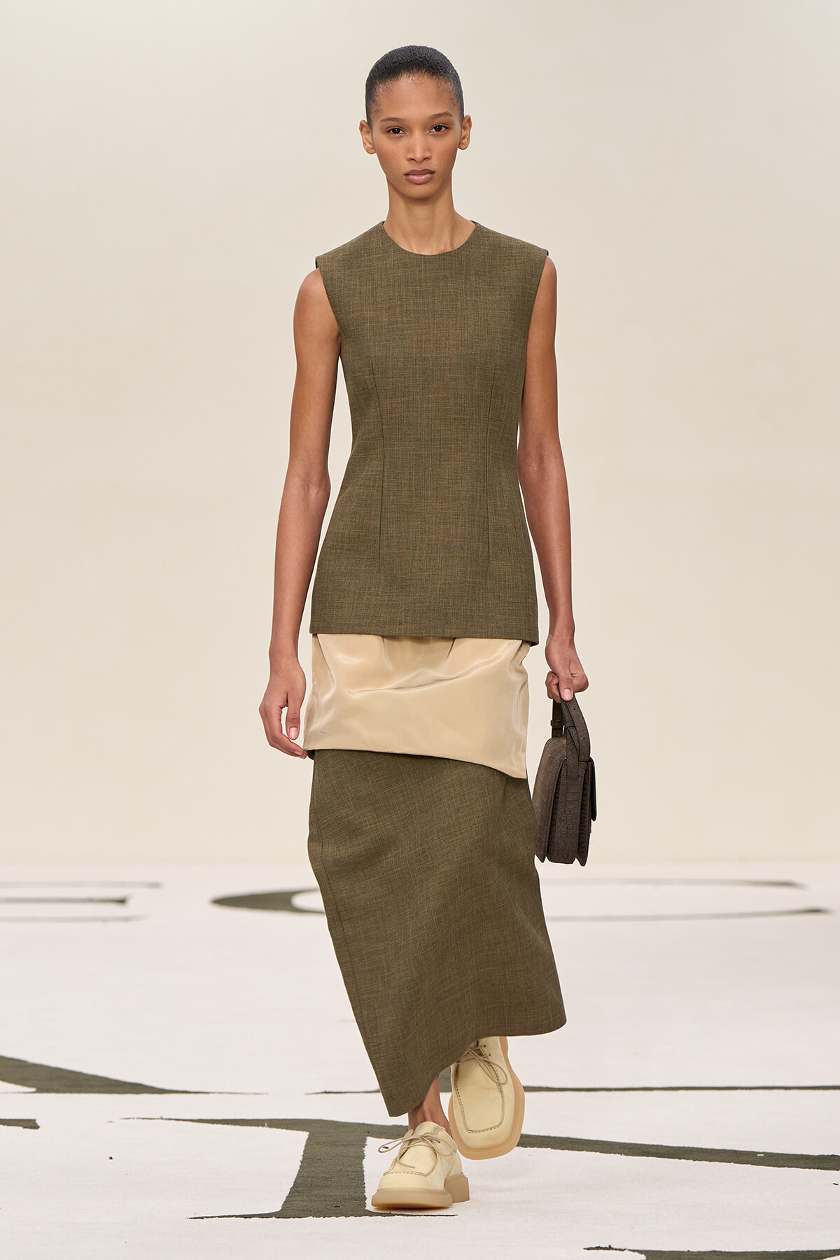 Model wearing an olive-green sleeveless top and long skirt with a beige satin waist panel, carrying a small brown handbag on a runway.