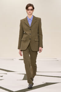 Model walking a runway in an olive brown tailored pantsuit with a light blue shirt and narrow sunglasses.