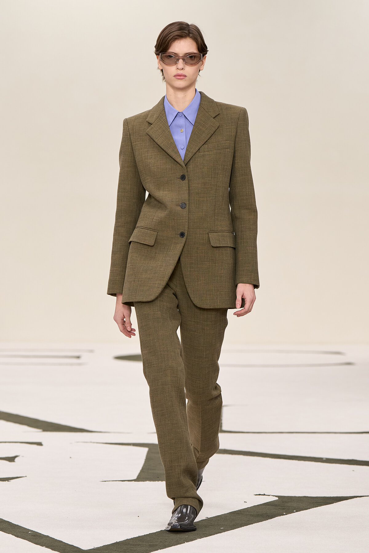 Model walking a runway in an olive brown tailored pantsuit with a light blue shirt and narrow sunglasses.