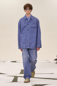 Male model in an oversized blue denim shirt with matching wide-leg jeans, sunglasses, walking on a runway.