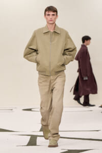 Front-view of a male model walking a runway in a beige zip jacket and matching trousers with hands in pockets.