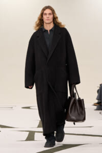 Male model walking a fashion show in a long black wool coat, carrying a large dark brown leather tote bag.