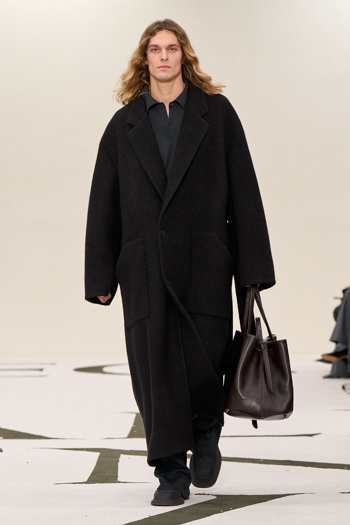 Male model walking a fashion show in a long black wool coat, carrying a large dark brown leather tote bag.