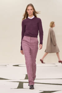 Female model walks a runway wearing a wine-colored sweater and pink plaid trousers, black ankle boots.