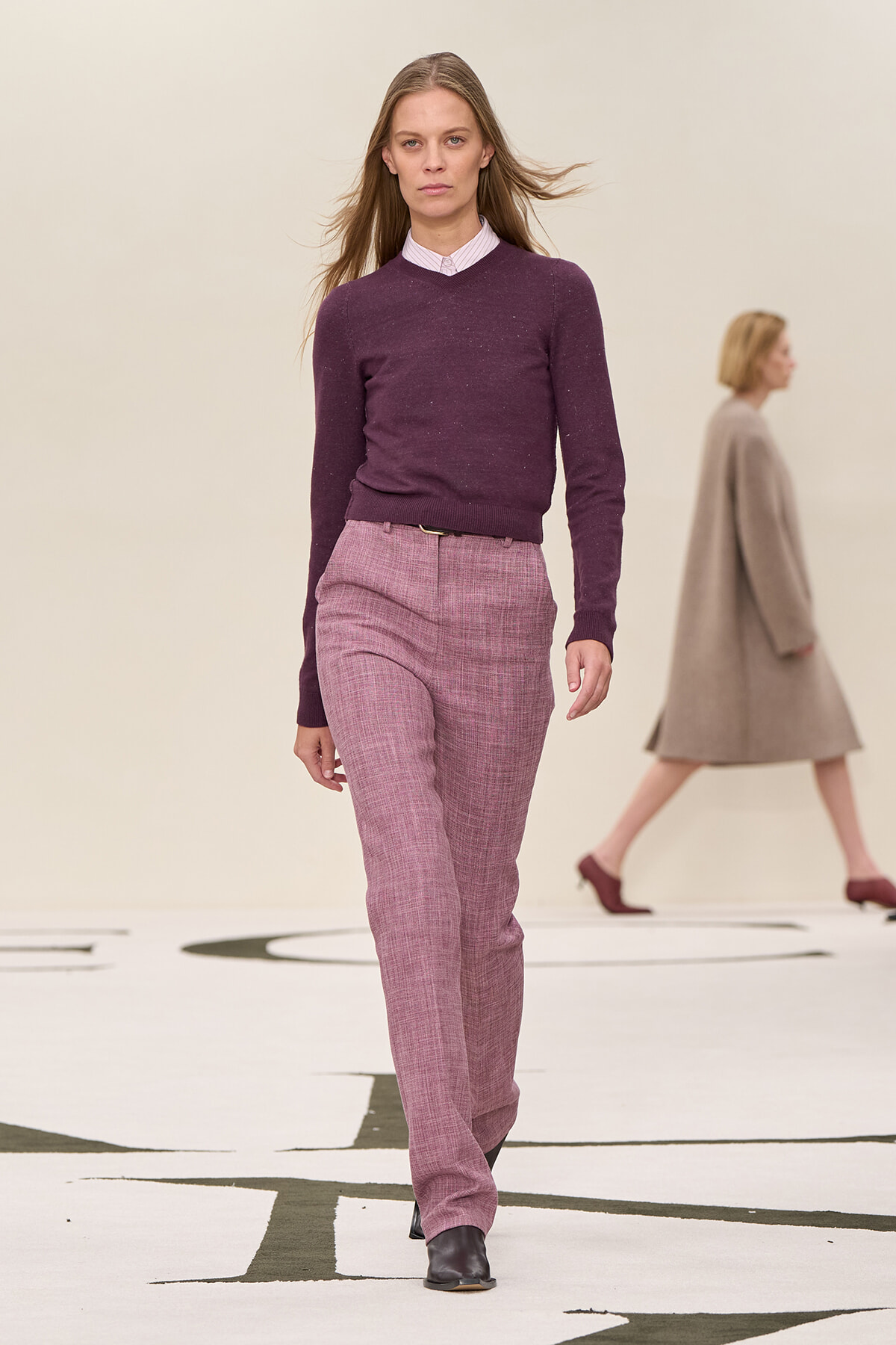 Female model walks a runway wearing a wine-colored sweater and pink plaid trousers, black ankle boots.
