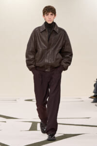 Male model walks the runway in a brown leather bomber jacket, black turtleneck, and dark trousers.