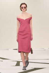Model walking a runway in a dusty pink off-shoulder midi dress, paired with gray heeled mules, round sunglasses, and a small black pendant bag on a thin strap.