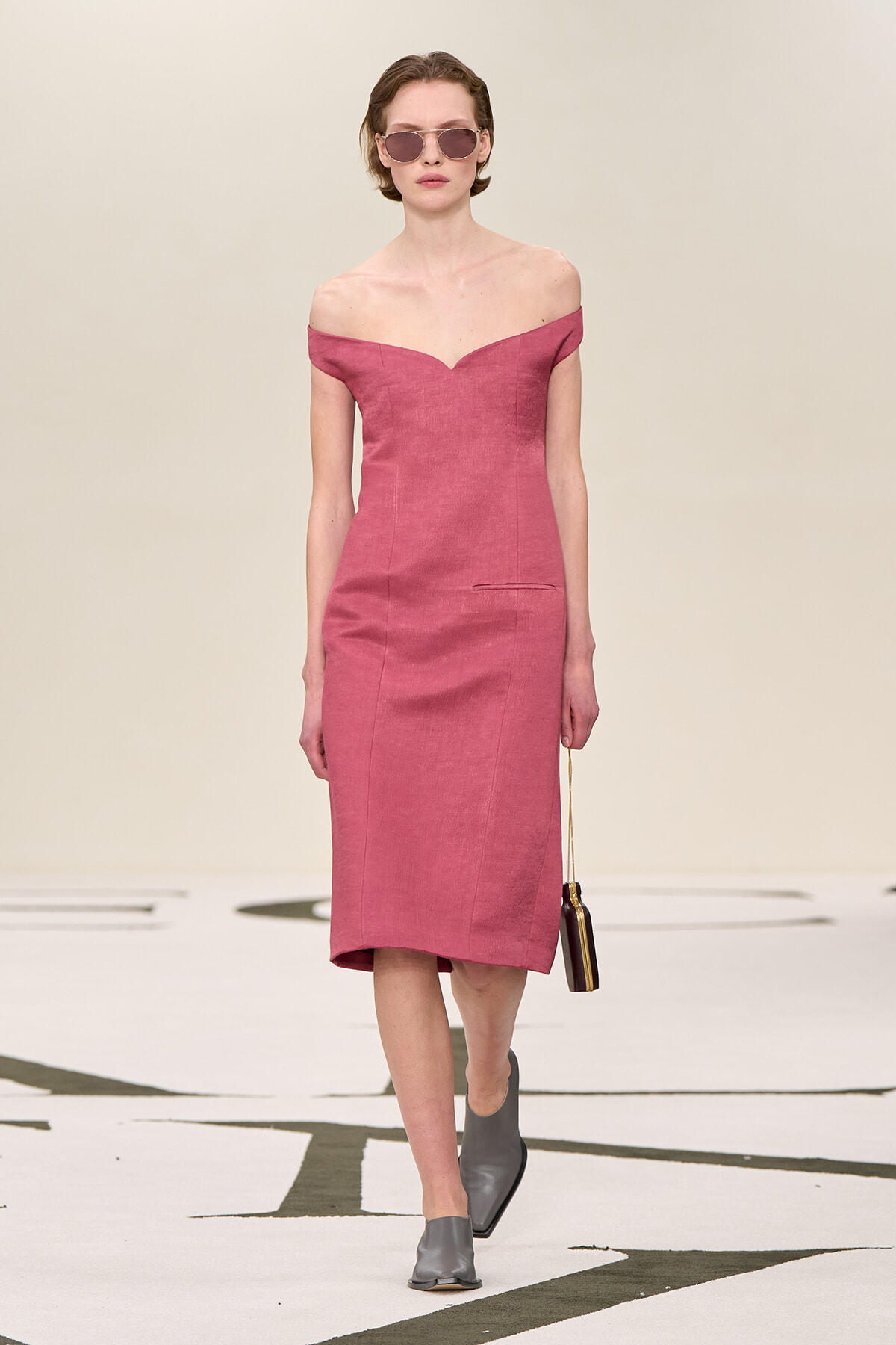 Model walking a runway in a dusty pink off-shoulder midi dress, paired with gray heeled mules, round sunglasses, and a small black pendant bag on a thin strap.