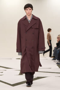 Male model walks the runway wearing a long burgundy double-breasted trench coat over a gray-cuffed shirt.