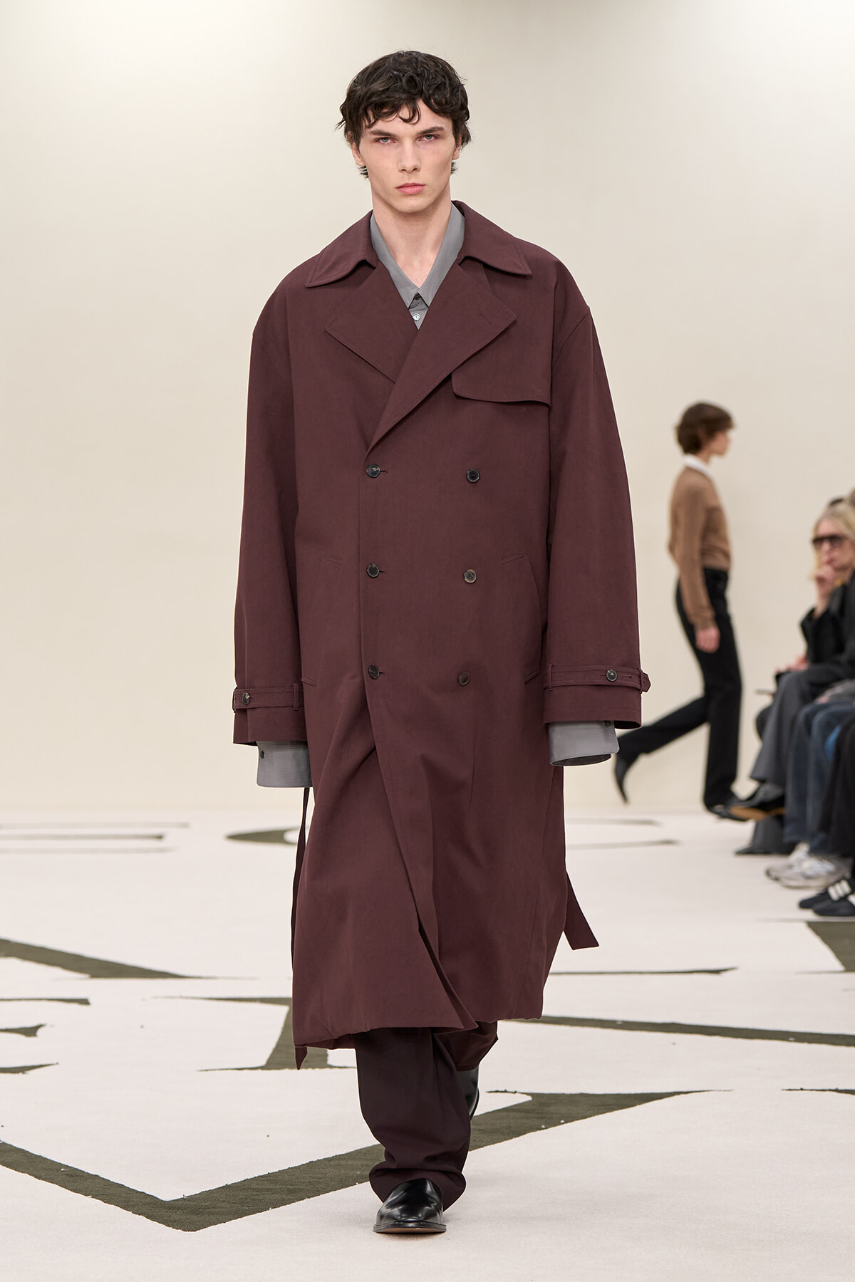 Male model walks the runway wearing a long burgundy double-breasted trench coat over a gray-cuffed shirt.