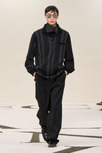 Male model walks a runway in a dark striped short-sleeve knit polo, black pants, and black sunglasses, minimalist styling.