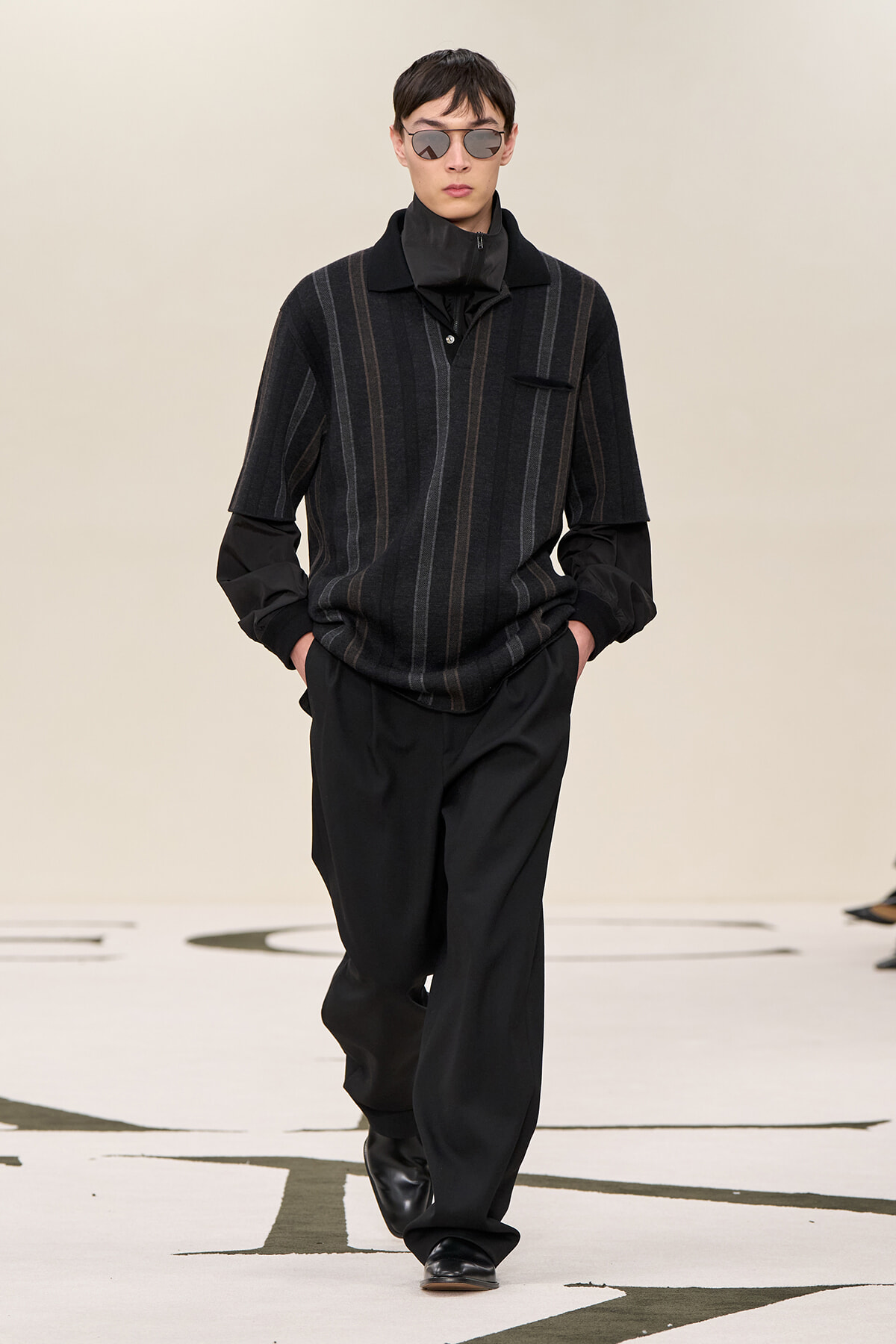 Male model walks a runway in a dark striped short-sleeve knit polo, black pants, and black sunglasses, minimalist styling.