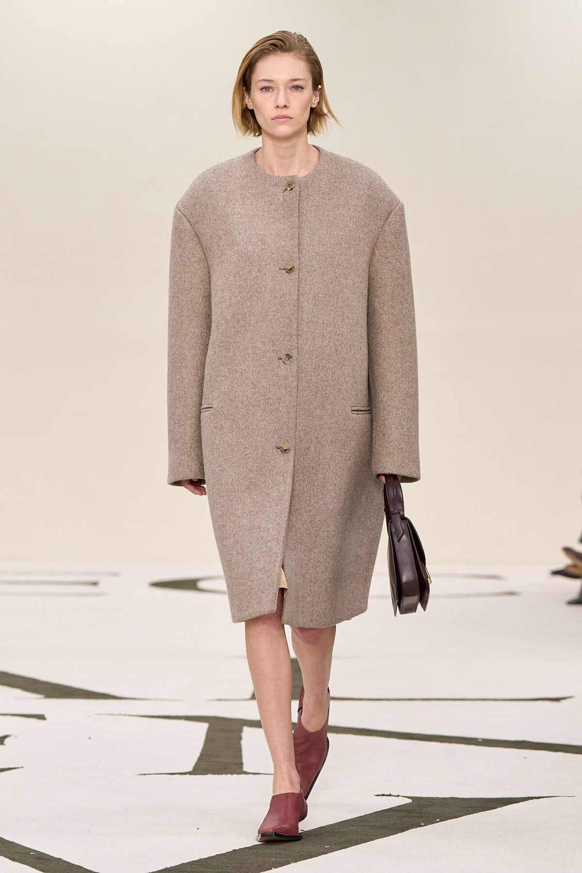 Model walking a fashion runway in a knee-length beige wool coat, holding a dark handbag and wearing burgundy shoes