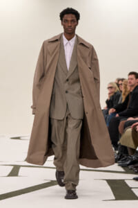 Male model walks a runway wearing a beige trench coat over a light gray suit and pink shirt, with spectators in the background.