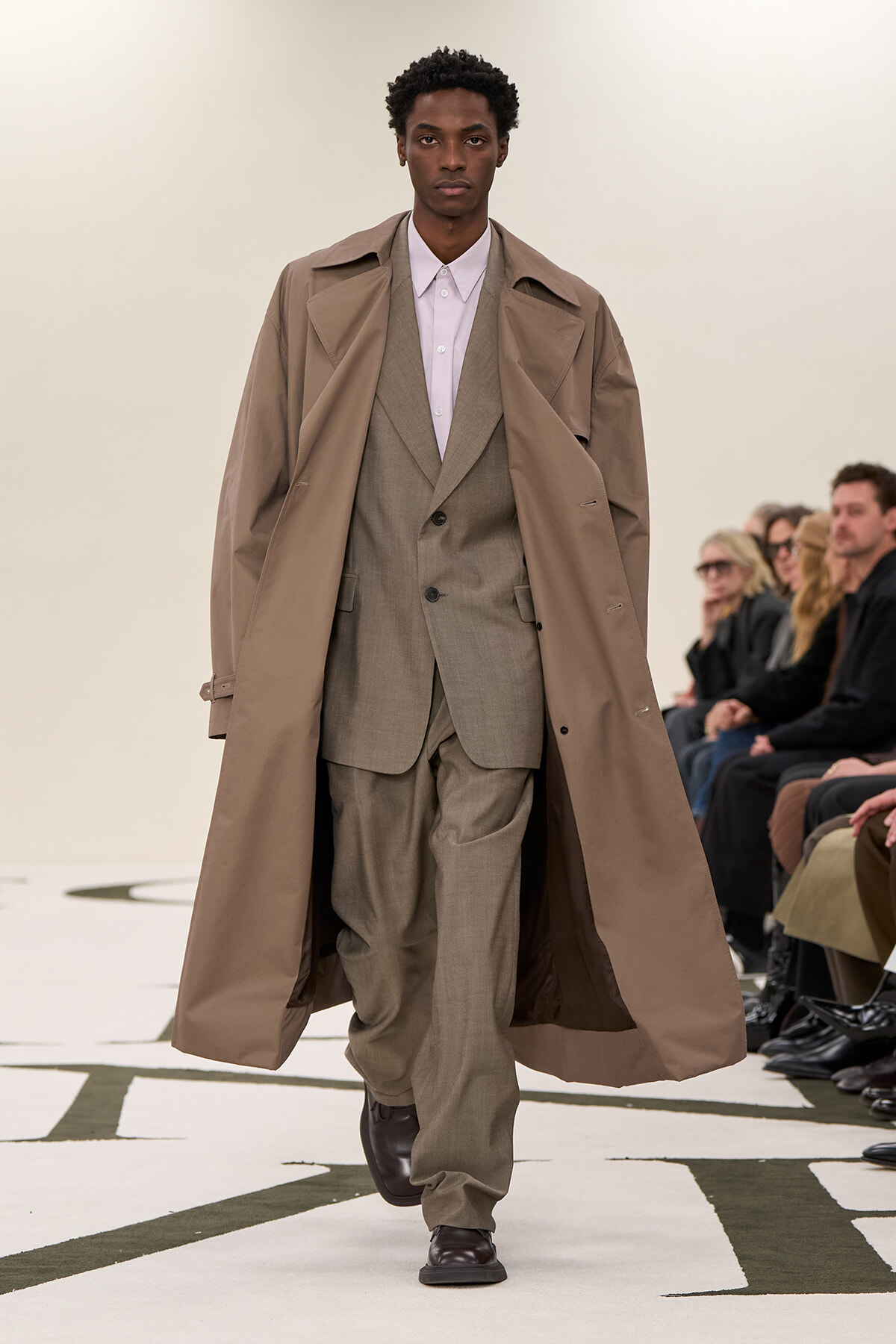 Male model walks a runway wearing a beige trench coat over a light gray suit and pink shirt, with spectators in the background.