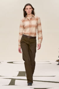 Model walking a runway in a beige plaid button-down shirt tucked into dark brown trousers with a brown belt in a neutral-toned look.