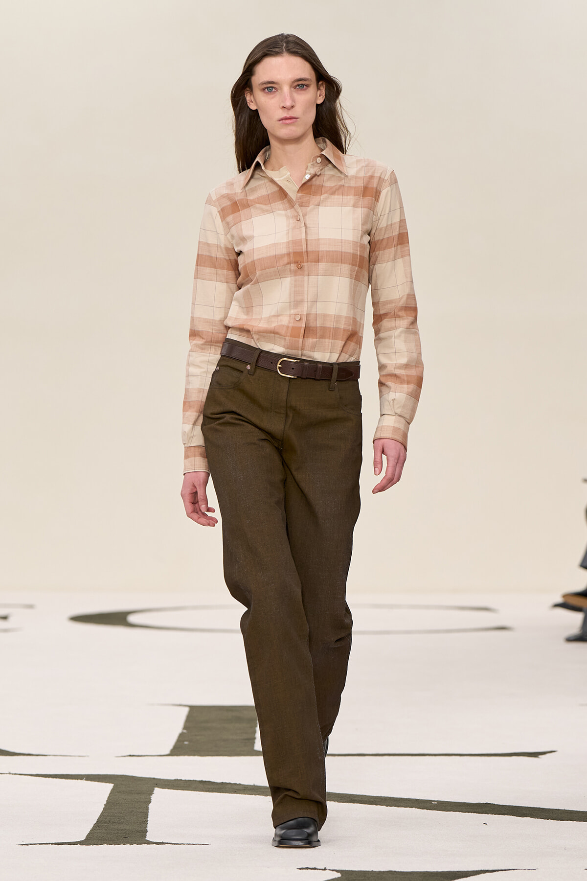 Model walking a runway in a beige plaid button-down shirt tucked into dark brown trousers with a brown belt in a neutral-toned look.