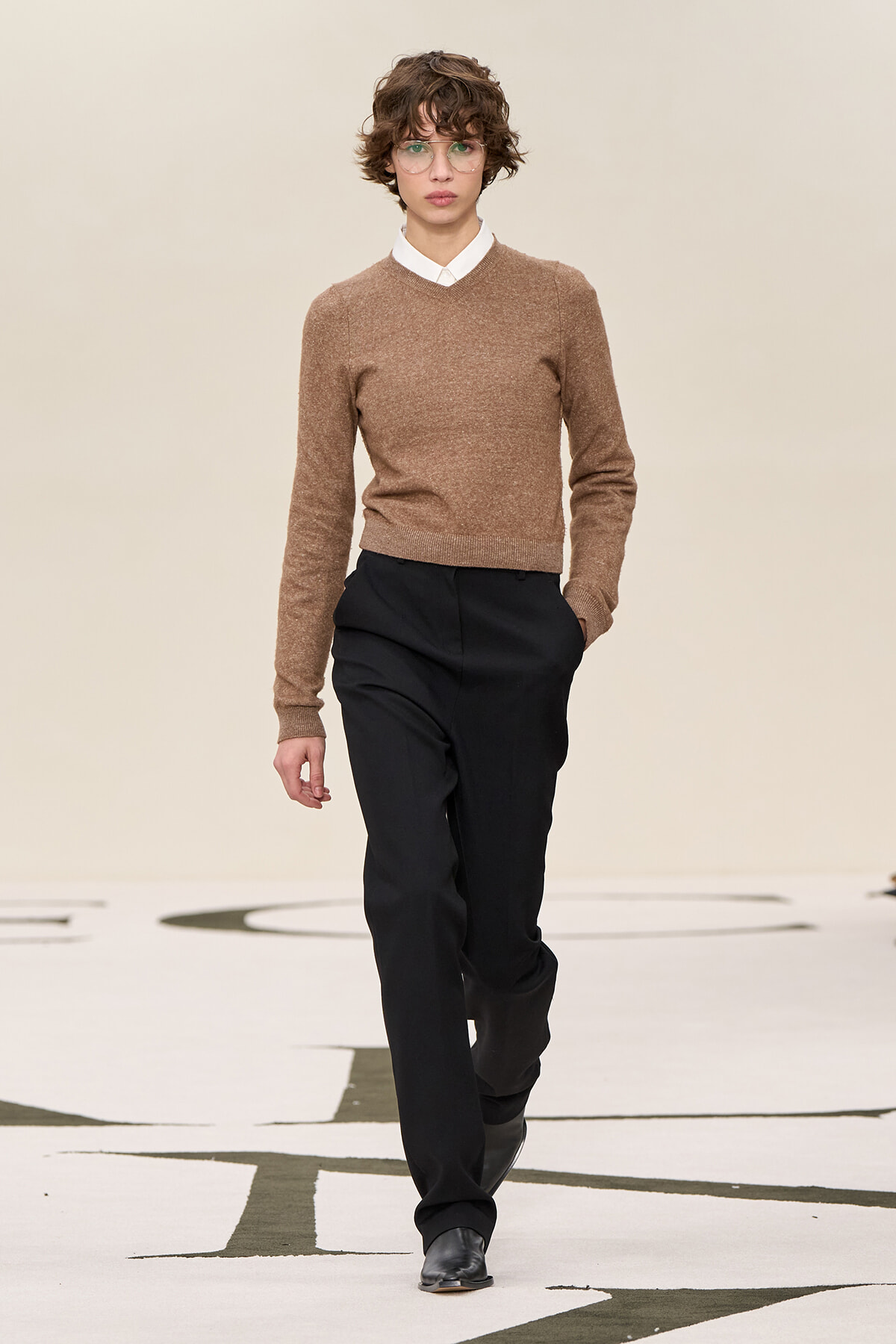 Model walks the runway in a brown knit sweater over a white collared shirt with black trousers and black shoes.